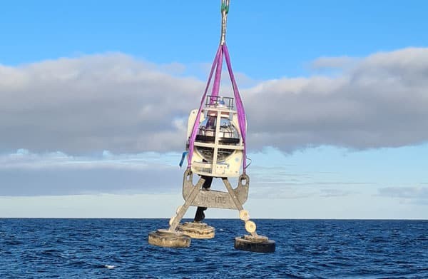 A white, spherical scientific instrument with three dark weights suspended by purple straps over a blue ocean.