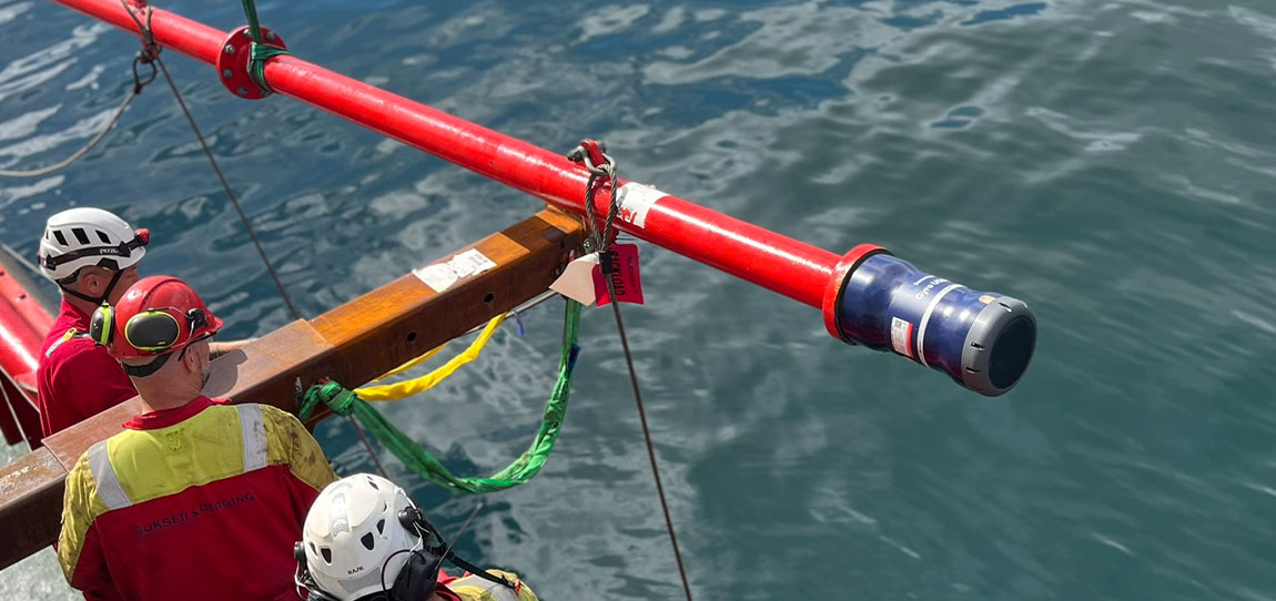 Three workers in safety gear oversee a red pipe with a blue and gray cylindrical device attached, extending over calm blue water during a marine operation.