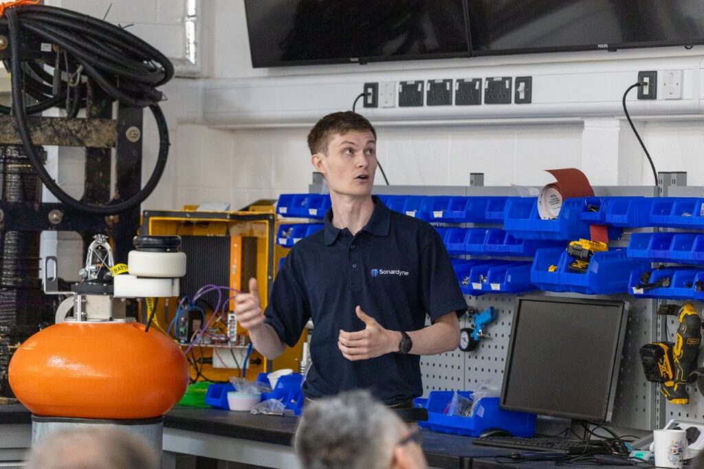A man in a Sonardyne polo shirt speaks in a workshop, gesturing towards an orange spherical device and a wall of blue storage bins.