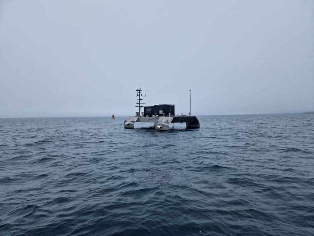 A dark gray autonomous catamaran vessel with 'AQUA' written on its side floats on dark, choppy water under an overcast sky, with a yellow buoy in the distance.
