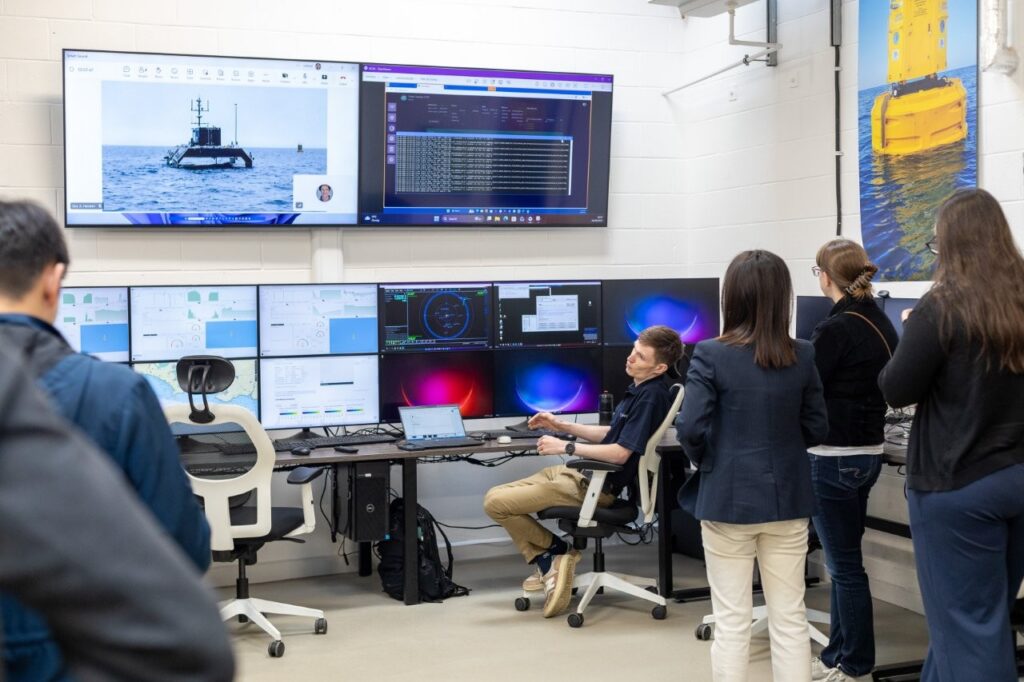 People in a control room looking at multiple computer screens displaying data, maps, and an unmanned surface vessel.