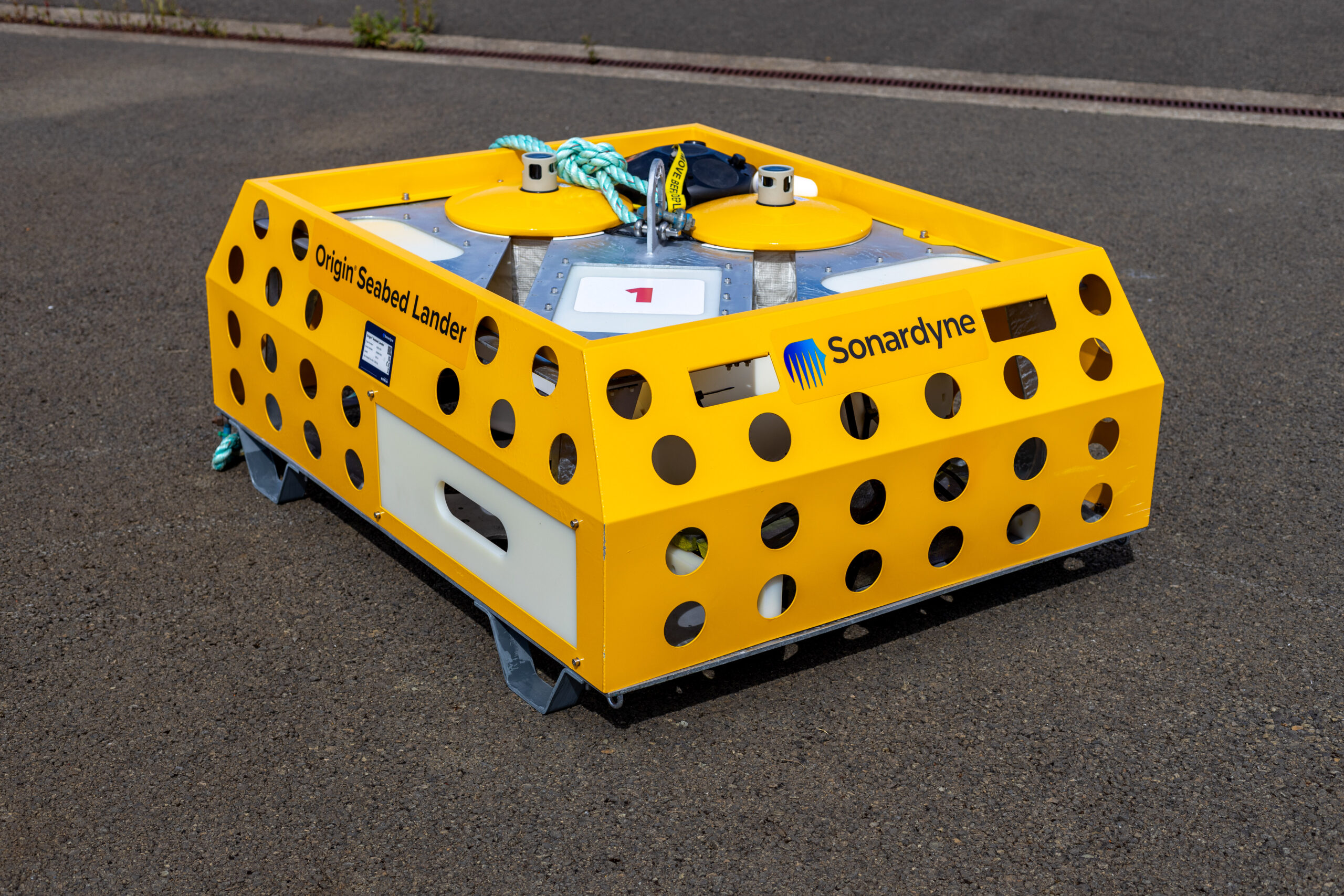 A bright yellow Origin Seabed Lander by Sonardyne, a rectangular device with a perforated cage-like frame, sits on dark asphalt.