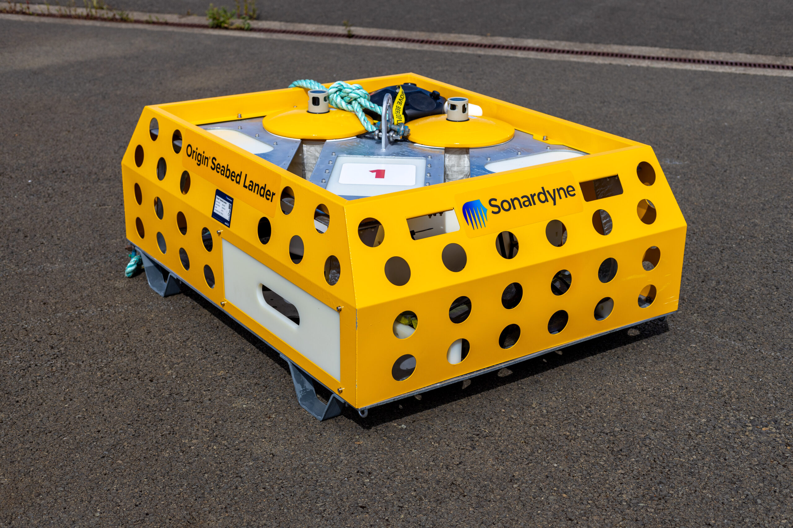 A bright yellow Origin Seabed Lander by Sonardyne, a rectangular device with a perforated cage-like frame, sits on dark asphalt.