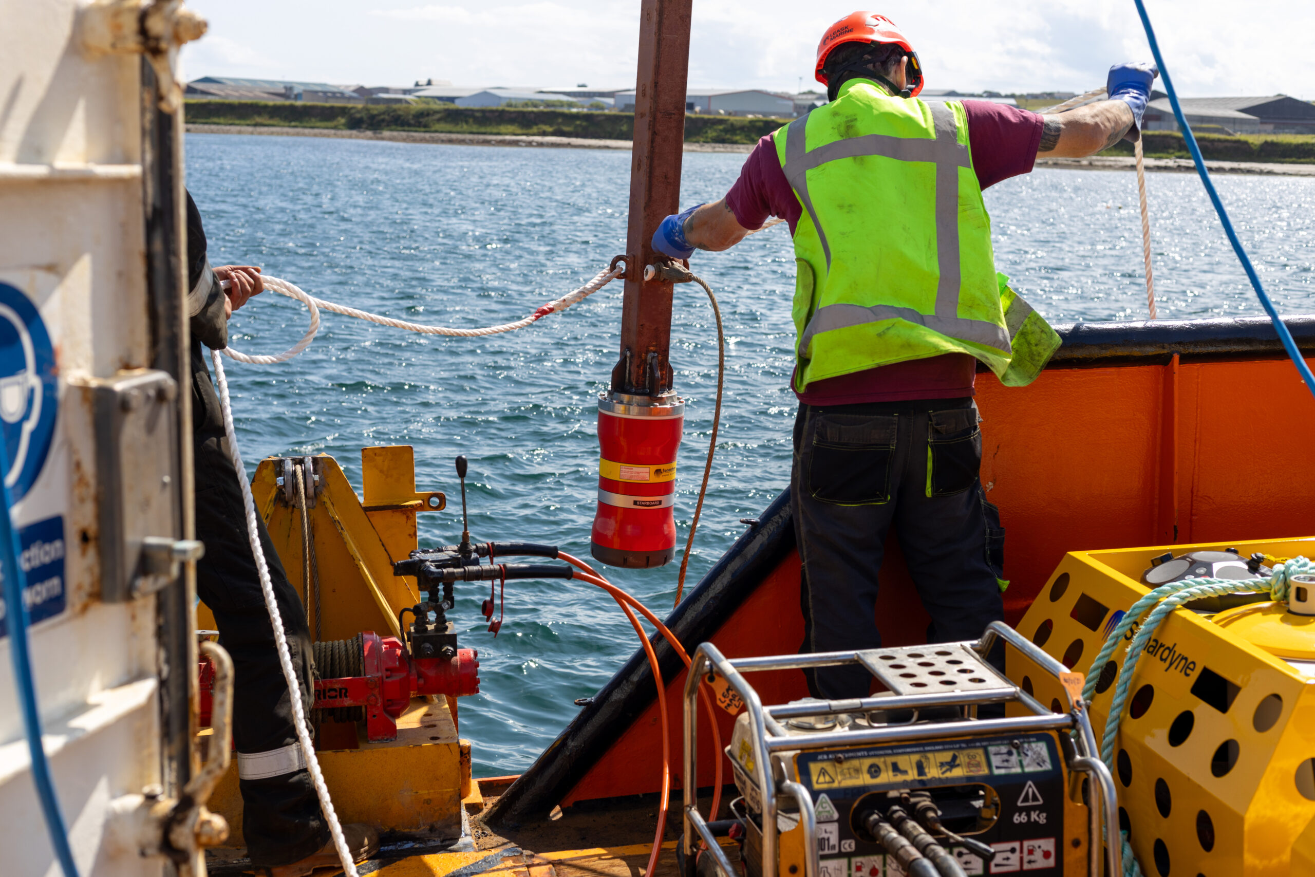 A marine worker in a hard hat and high-visibility vest on a boat, handling a rope attached to a red cylindrical device suspended over blue water.