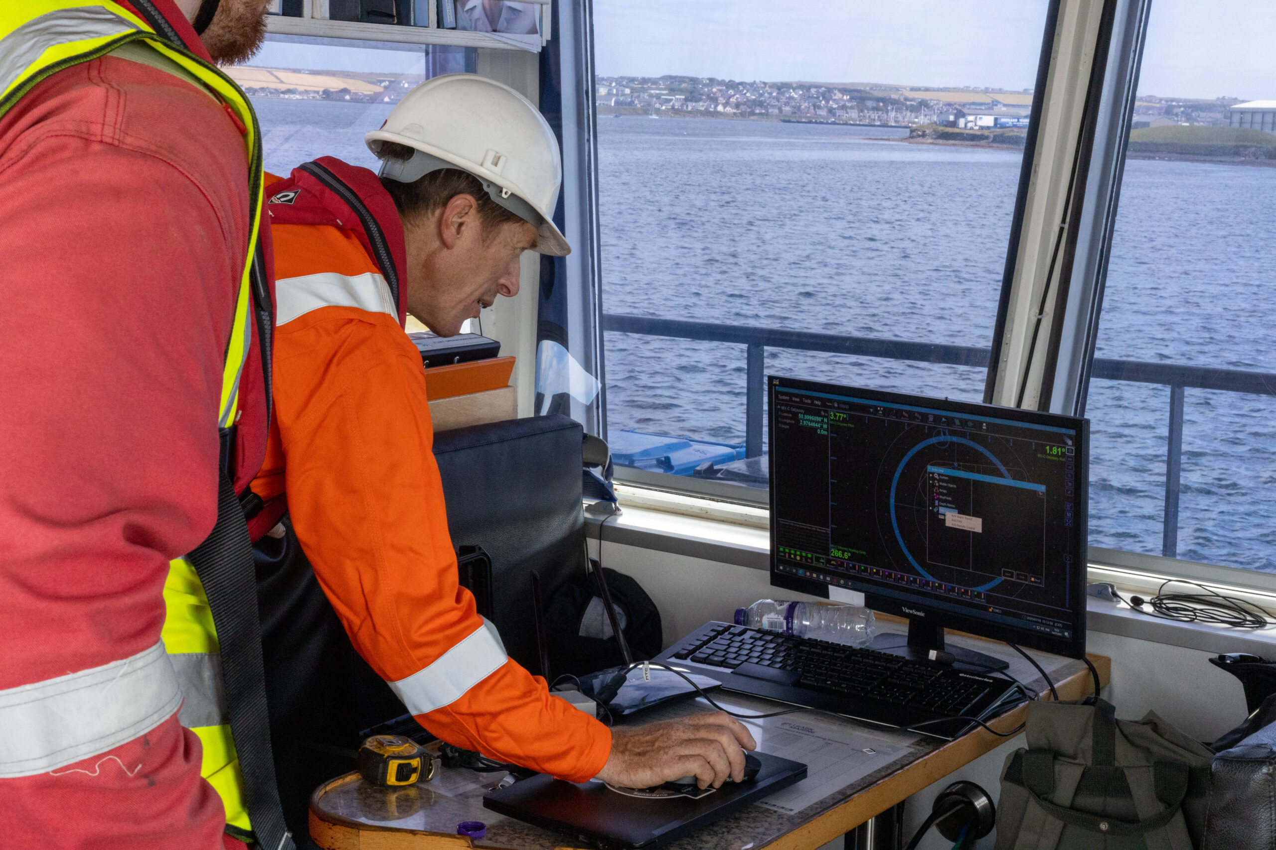 A person in an orange safety suit and white hard hat uses a computer with navigation software on a ship's bridge, with a view of the sea and a distant town.