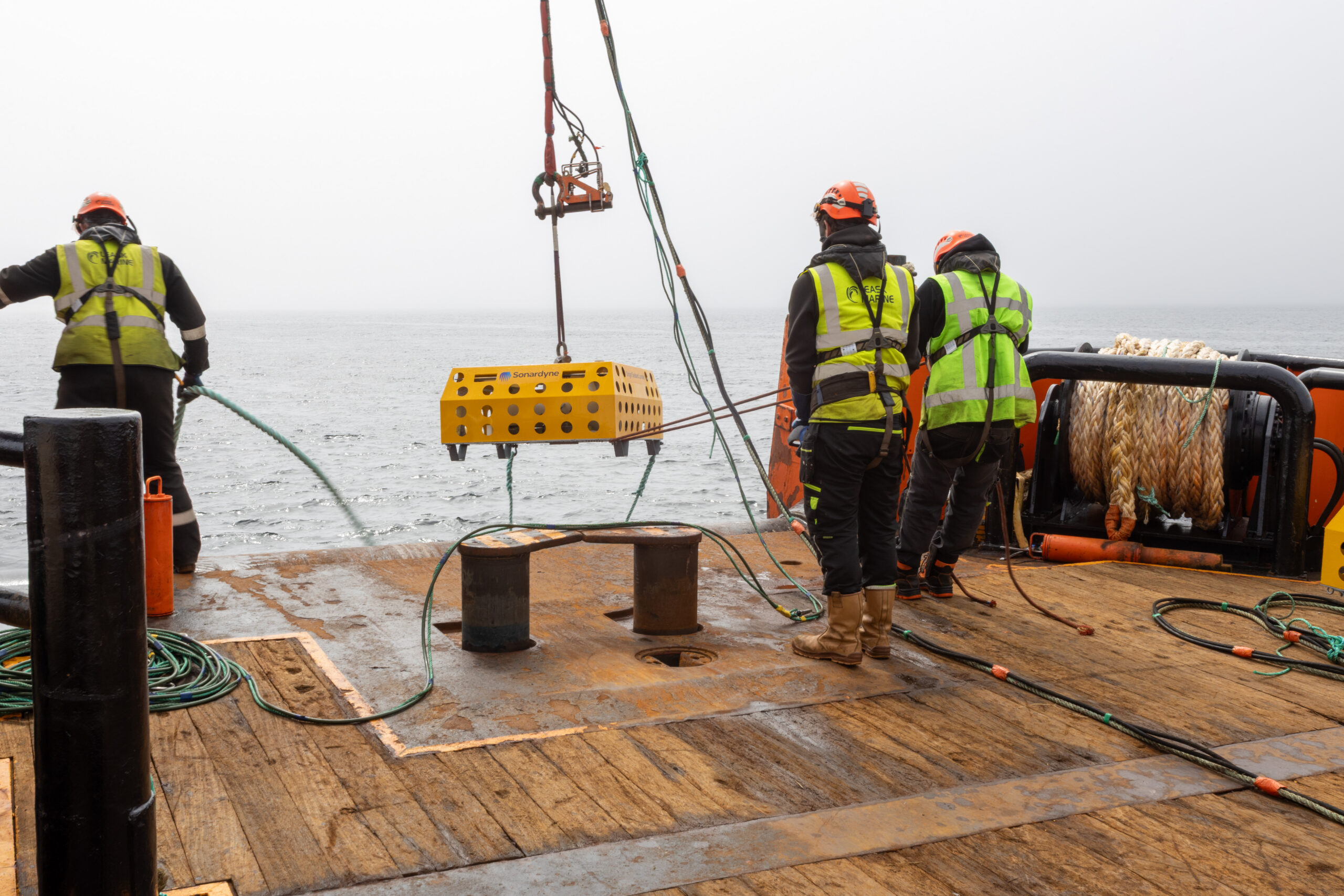 Three crew members in safety gear on a boat deck, with a yellow Sonardyne cage suspended above the deck, overlooking a calm, grey sea.