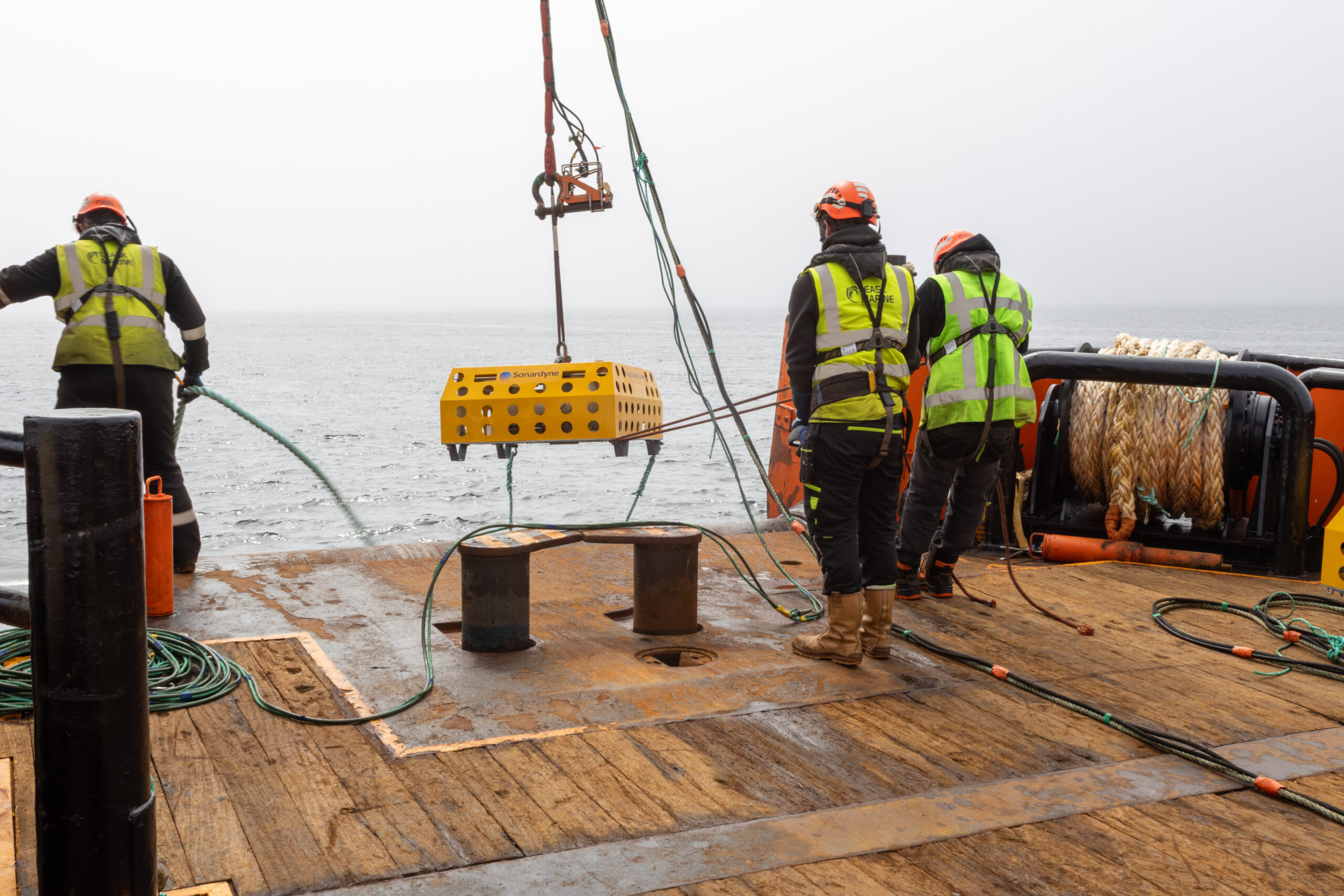 Three crew members in safety gear on a boat deck, with a yellow Sonardyne cage suspended above the deck, overlooking a calm, grey sea.