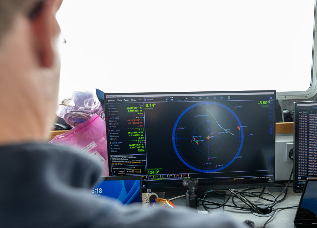 A person views two computer monitors displaying marine navigation and communication software. The left screen shows a sonar tracking interface with ship positions and beacon data, while the right screen displays a communication log. A laptop, camera, and cables are on the desk.