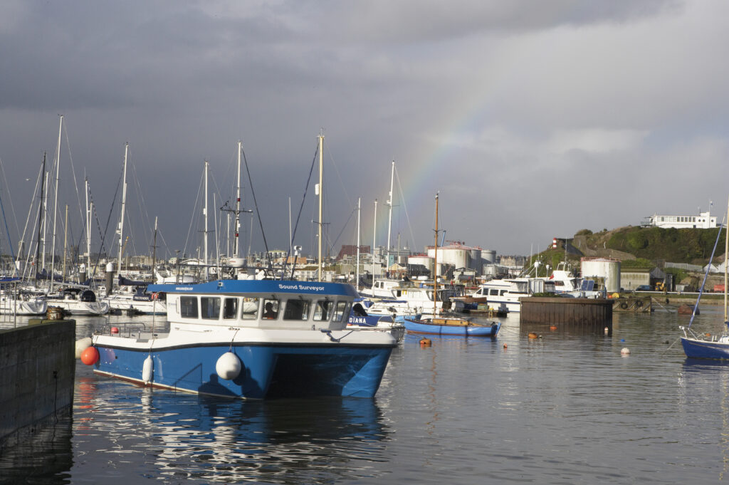 A blue and white boat named 'Sound Surveyor' in a marina with many sailboats, under a sky with a visible rainbow.