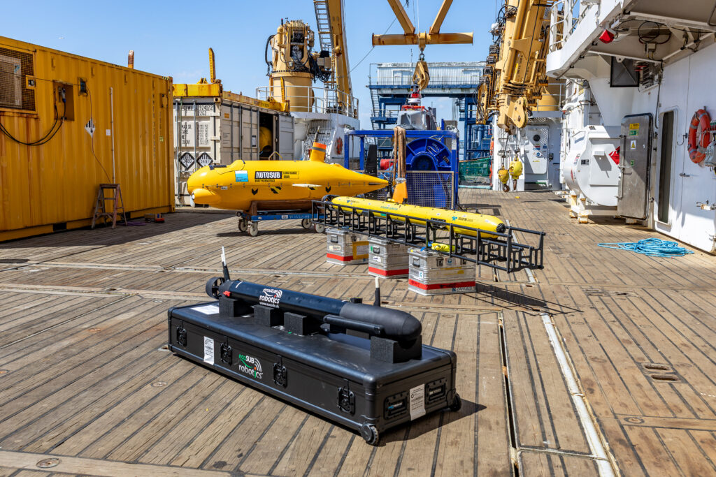 Three autonomous underwater vehicles (AUVs) of varying sizes and colors, along with shipping containers and cranes, on the wooden deck of a ship under a clear blue sky.