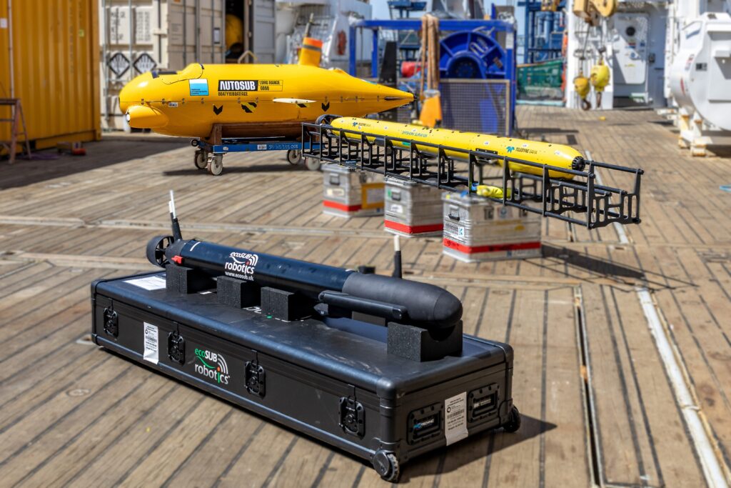 Three autonomous underwater vehicles (AUVs) – one large yellow, one medium yellow, and one small black – on the wooden deck of a ship.