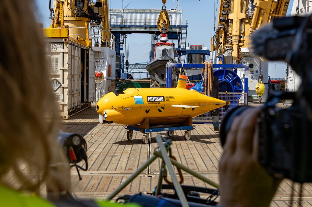 A bright yellow autonomous underwater vehicle (AUV) named "Autosub Boaty McBoatface" sits on a wheeled trolley on the wooden deck of a ship, surrounded by cranes and shipping containers.