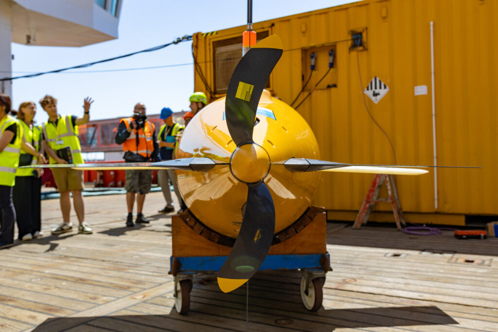 A large yellow autonomous underwater vehicle with a four-bladed propeller on a wheeled cart on a wooden deck, with people in high-visibility vests and a yellow container in the background.