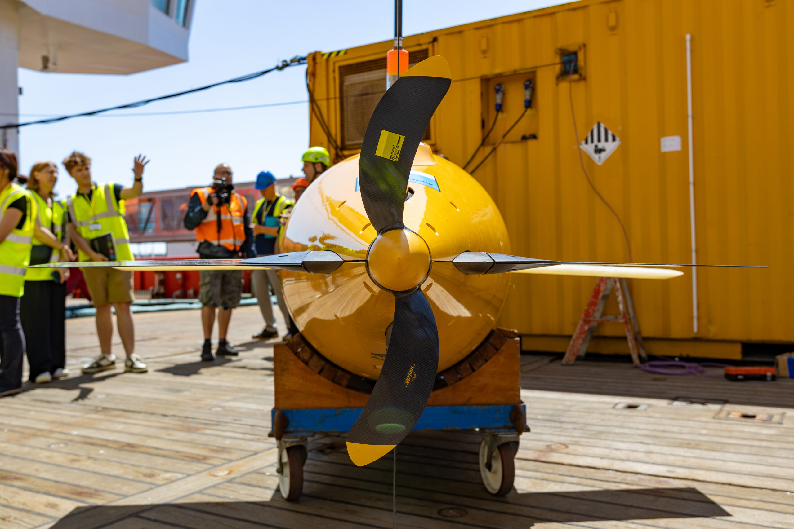 A large yellow autonomous underwater vehicle with a four-bladed propeller on a wheeled cart on a wooden deck, with people in high-visibility vests and a yellow container in the background.