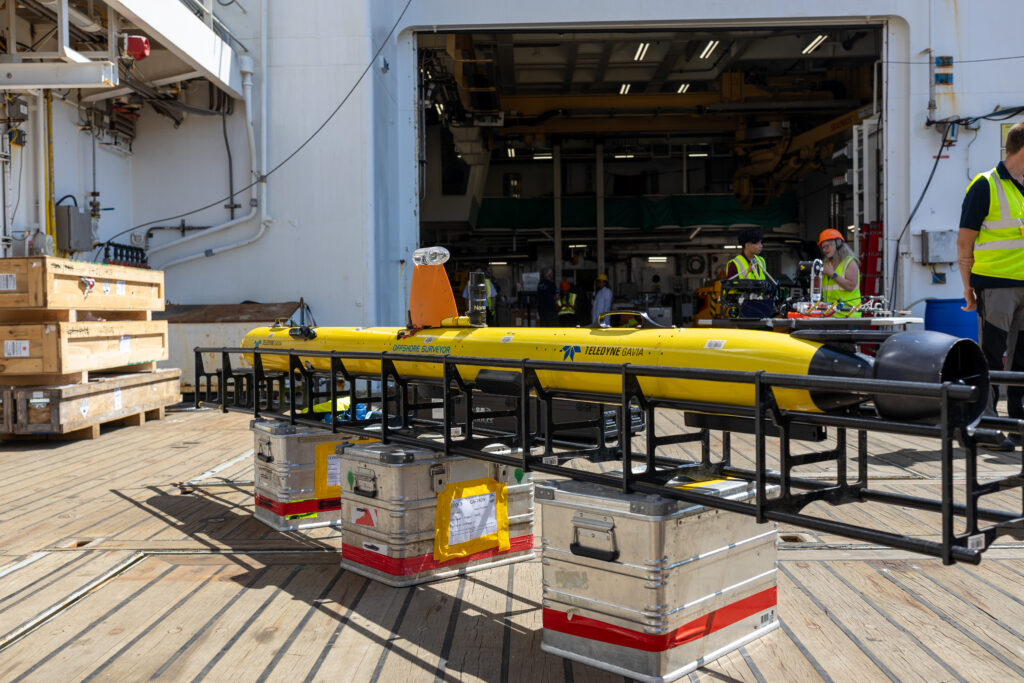 A bright yellow Teledyne Gavia 'Offshore Surveyor' AUV on a ship's wooden deck, supported by a black frame and silver cases, with crew members and industrial equipment visible in the background.