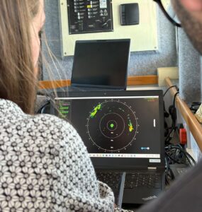 Over-the-shoulder view of a person looking at a laptop screen displaying a circular radar image with green, yellow, and red clusters. Another laptop and a control panel are in the background.
