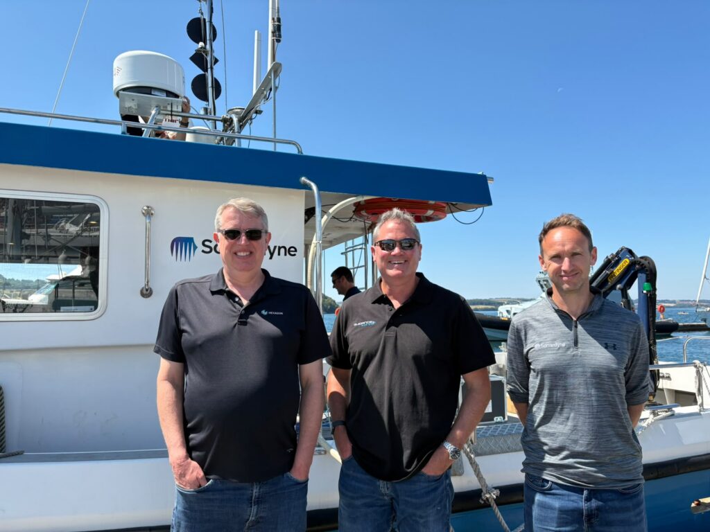 Three smiling men, wearing polo shirts and jeans, stand in front of a white and blue Sonardyne boat on a sunny day.