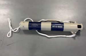 A beige cylindrical Sonardyne RT 6-1000 acoustic transponder with blue labels and white rope attached, lying on a gray surface.
