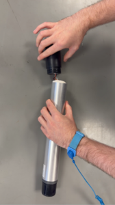 A person's hands assembling a silver and black cylindrical device on a gray surface. A blue anti-static wrist strap is visible on the right wrist.