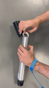 Close-up of hands assembling a silver and black cylindrical device, placing a white ring into the opening. A blue ESD wrist strap is visible on the left wrist.