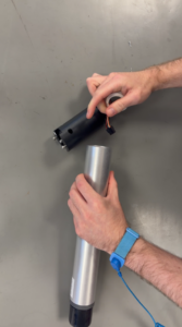 Two hands working with cylindrical components. The right hand holds a black cylinder with wires, and the left hand holds a silver cylinder. A blue anti-static wrist strap is on the left wrist.