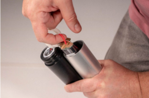 Hands holding two cylindrical devices, one black and one silver, with internal wiring and circuit board visible.