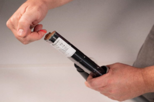 A person's hands pulling a red tab from the top of a black cylindrical object, which is partially inserted into a grey tube.