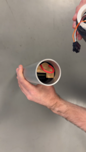 A person's hands assembling electronic components. A silver tube holds a brown component with a red wire, while a black connector with wires is held nearby.