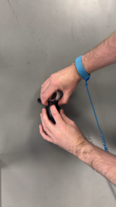 A person's hands, wearing a blue ESD wrist strap, manipulate a small dark cylindrical object with visible internal wiring against a gray background.