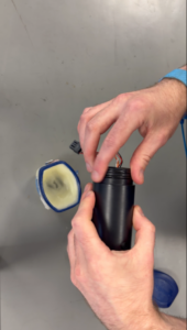 A person's hands inserting wires into a black cylinder. A container with a yellow substance and a blue lid are also on the gray surface.