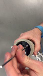 Close-up of hands using a screwdriver to work on a cylindrical electronic device with a beige casing and black interior. A blue wristband is visible on one wrist.