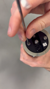 Close-up of hands using a hex screwdriver on a cylindrical device with a black top panel and a label '3'.