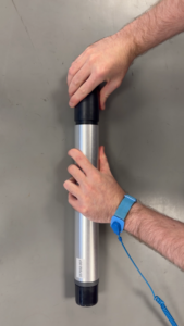 Two hands holding a silver cylindrical object with black caps, one hand wearing a blue anti-static wrist strap.
