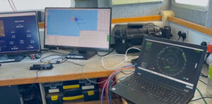 A workstation on a boat with three computer screens and a laptop displaying navigation, sonar, and radar data. Cables are visible on the wooden desk.