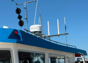 Upper part of a boat with a blue roof and white cabin, featuring a radar dome, multiple antennas, navigation signals, and a wind vane against a clear blue sky with a crescent moon.