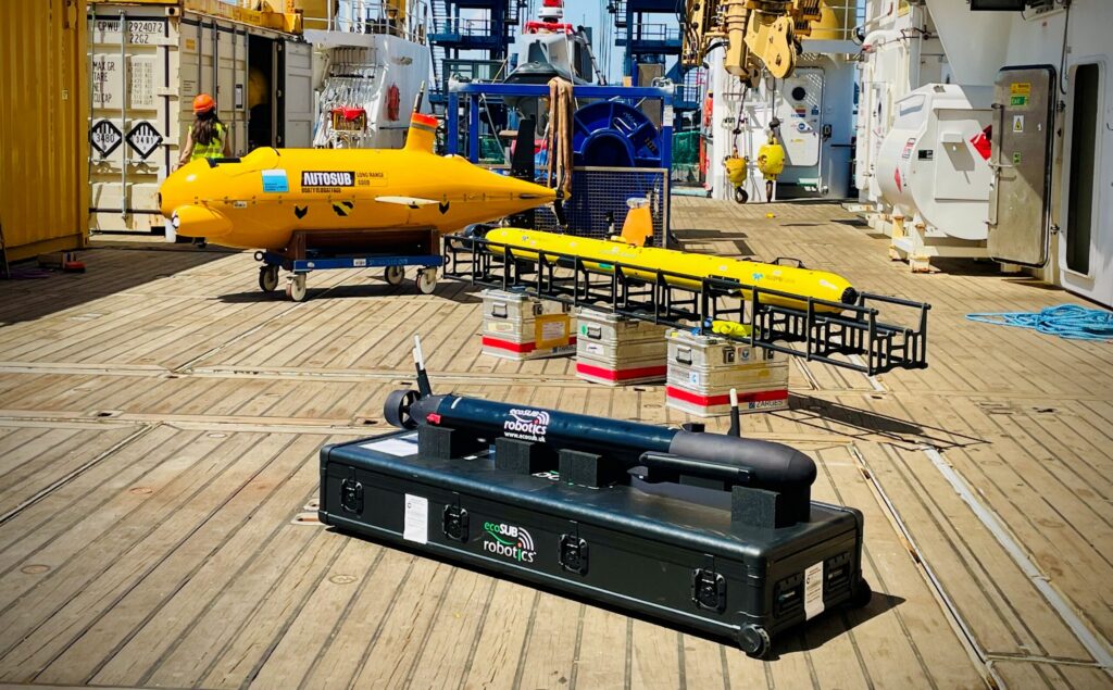Three autonomous underwater vehicles (AUVs) – a large yellow AUTOSUB, a long yellow Teledyne Gavia, and a black ecoSUB Robotics vehicle – displayed on the wooden deck of a ship with cranes and containers in the background.