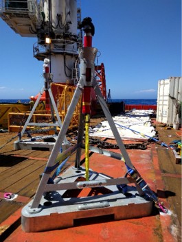 A large, silver triangular metal frame with a central red and silver cylinder on a ship's deck, with the ocean in the background.