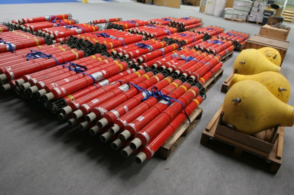 Red cylindrical equipment bundled on pallets next to large yellow pear-shaped buoys on pallets in a warehouse.