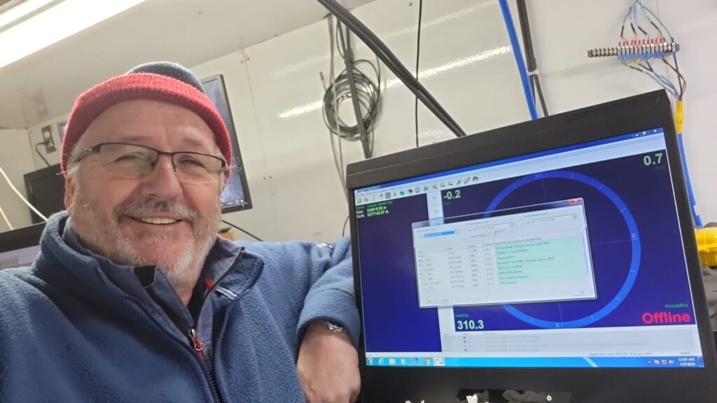 A man in a beanie and fleece jacket smiles next to a computer monitor displaying technical data, including 'Acoustics Offline' and a heading of 310.3.
