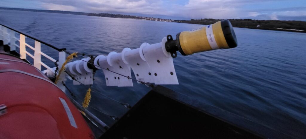 A yellow cylindrical sensor and white fin-like structures extend from the side of a boat over dark water, with a distant treelined shore and overcast sky.