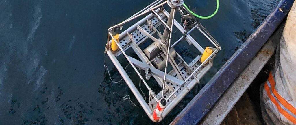 A metal scientific instrument with yellow and red sensors is suspended over dark blue ocean water from a boat.