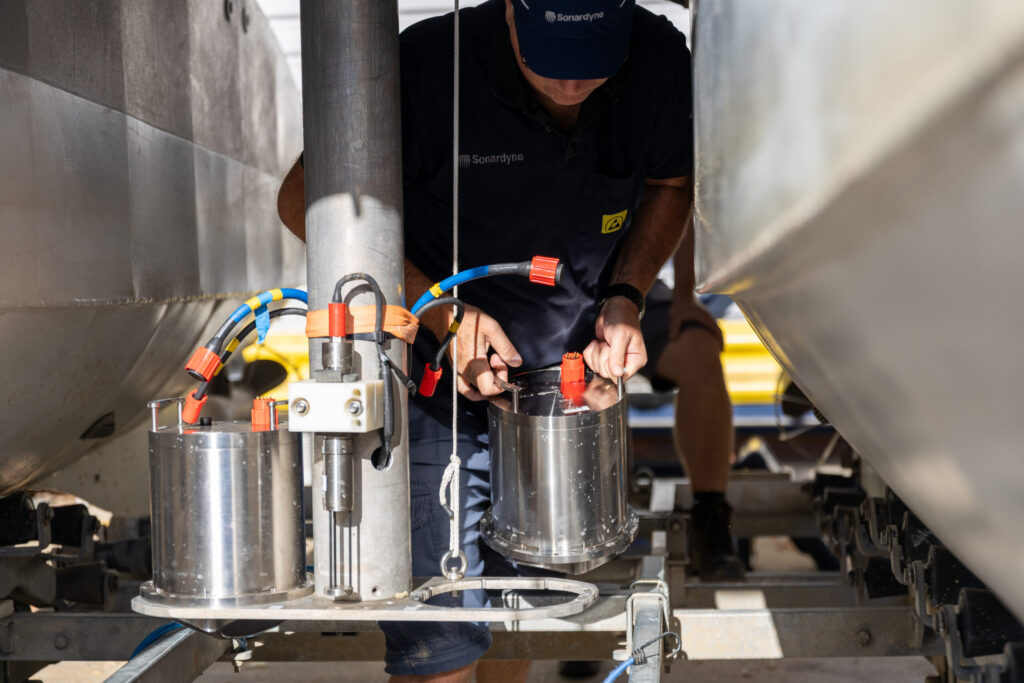 A Sonardyne technician in a blue uniform works on cylindrical marine sensors mounted on a metal frame, with boat hulls visible in the background.