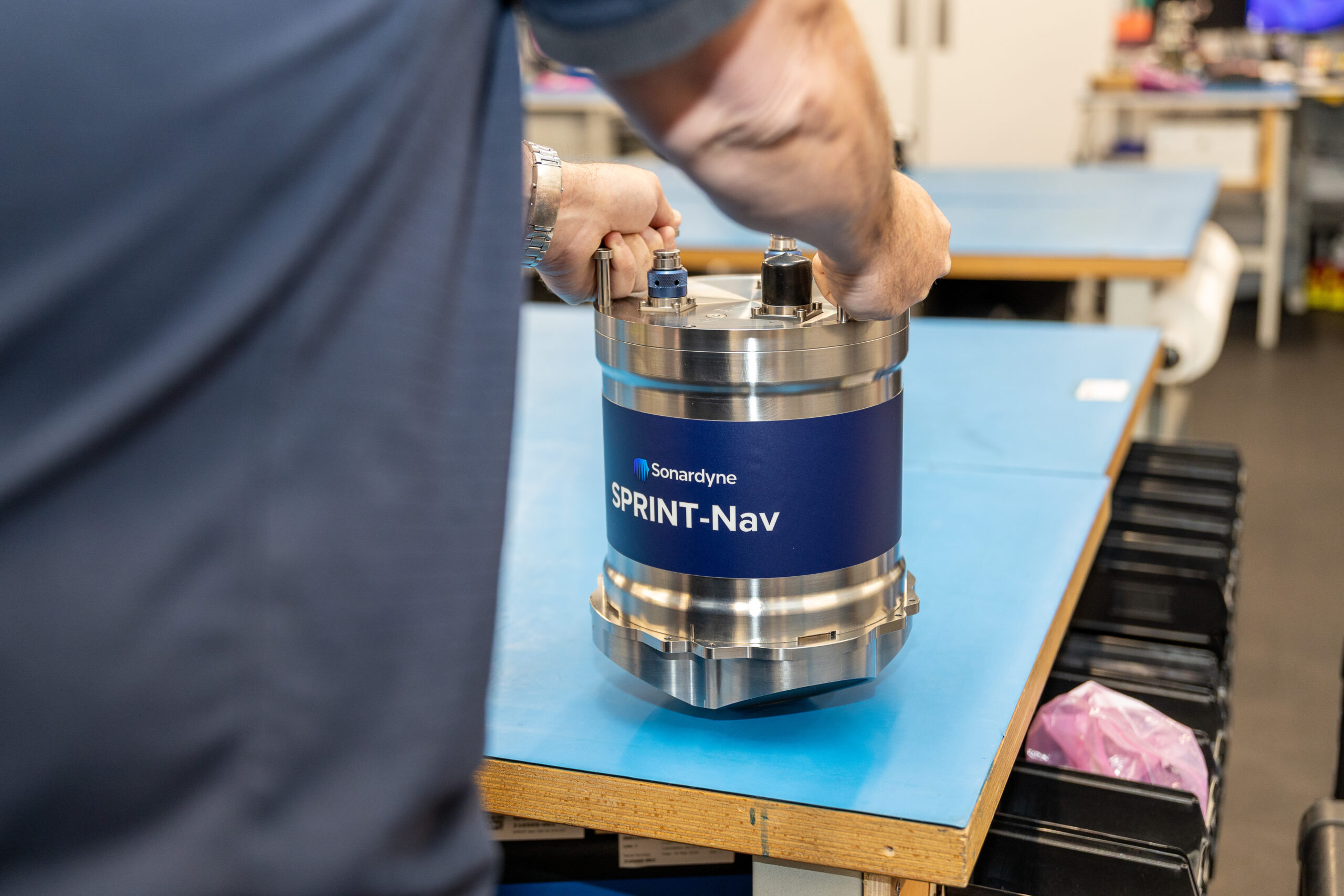 A person's hands assemble a cylindrical Sonardyne SPRINT-Nav device on a blue workbench.