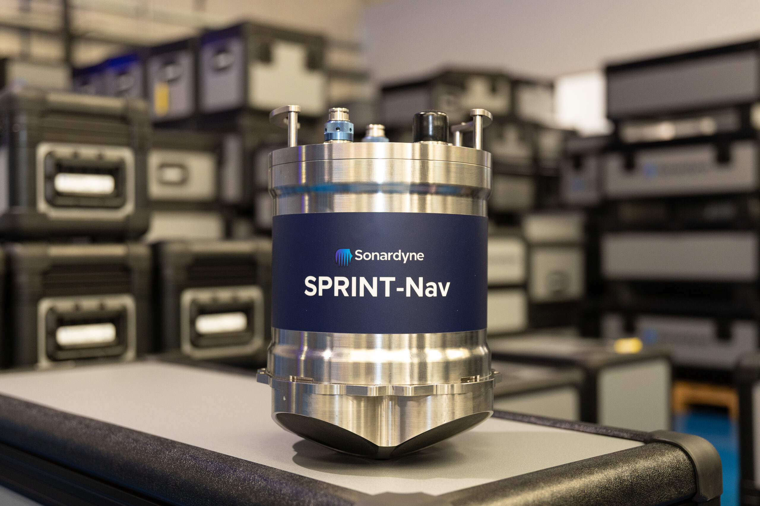 A cylindrical, silver-colored Sonardyne SPRINT-Nav device with a blue label, sitting on a grey surface with a blurred background of industrial cases.