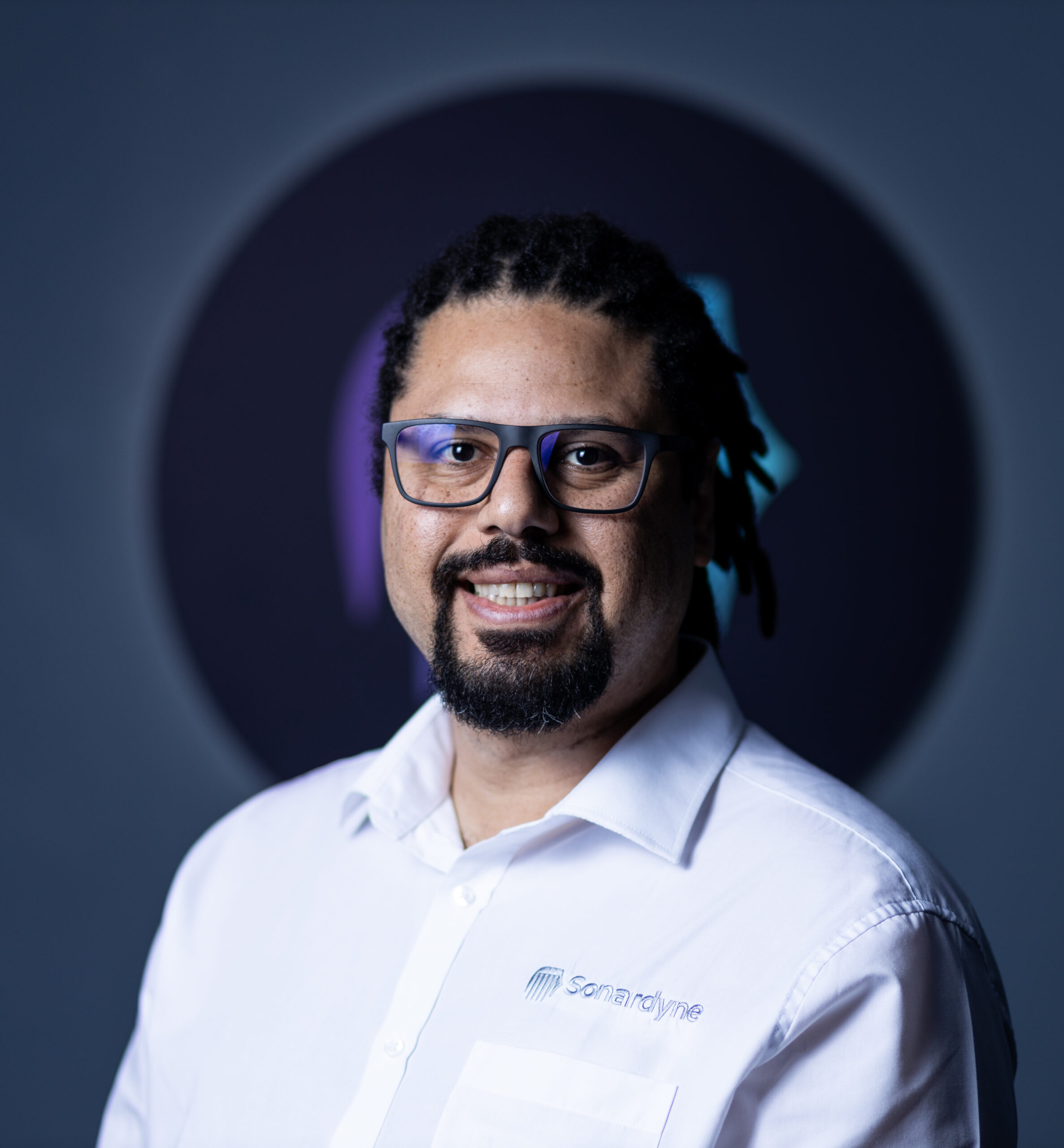 A man with dreadlocks, glasses, and a beard smiles, wearing a white shirt with the Sonardyne logo.