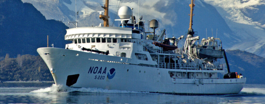 A white NOAA research ship with 'S 220' on its side moves through water, with towering snow-capped mountains in the background under a blue sky.