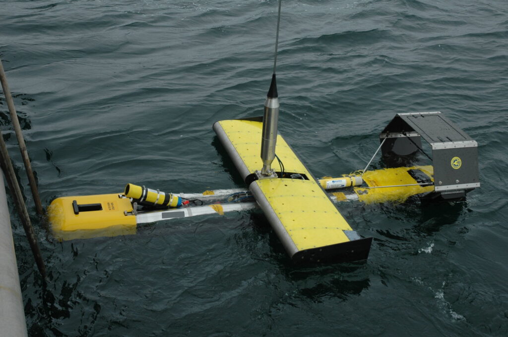 A yellow and silver autonomous ocean glider floats on dark, choppy water, with a central antenna and various sensors visible.
