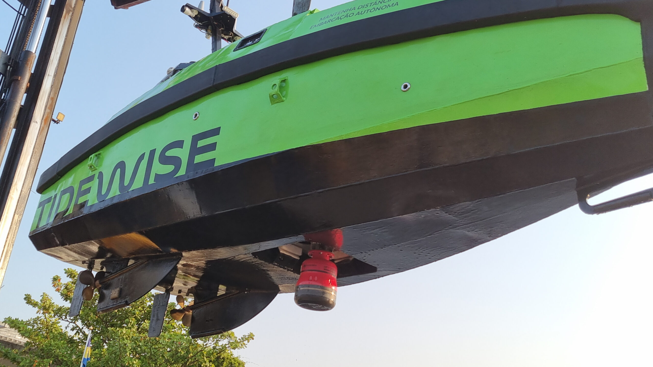 A green and black autonomous vessel named TIDEWISE is lifted against a blue sky, showing its propellers and a red sensor on the underside.
