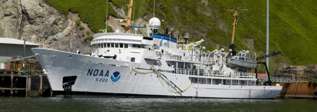 A white NOAA research vessel, S 220, docked in water with a steep green hillside in the background.