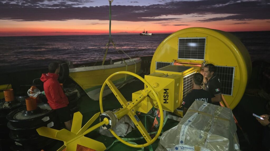 A vibrant sunset over the sea with a ship's deck in the foreground. Yellow marine equipment, including a large buoy with solar panels, is visible, along with crew members working. Another vessel is in the distance.